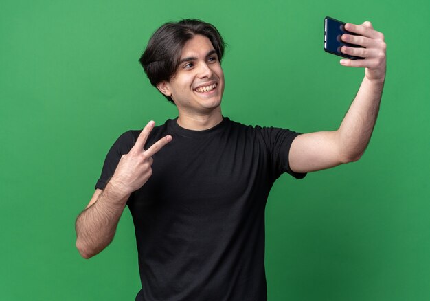 Smiling young handsome guy wearing black t-shirt take a selfie showing peace gesture isolated on green wall