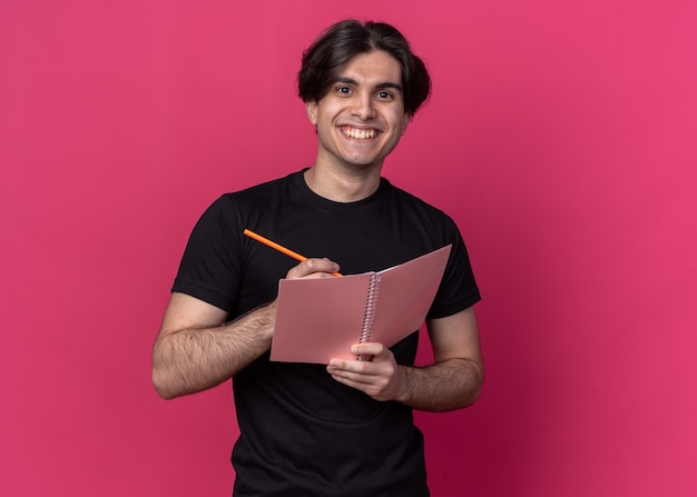 Smiling young handsome guy wearing black t-shirt writing something on notebook isolated on pink wall