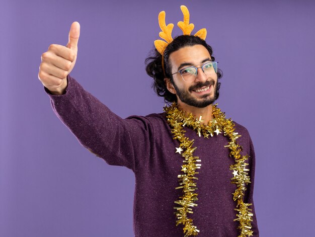 Smiling young handsome guy wearing christmas hair hoop with garland on neck showing thumb up isolated on blue background