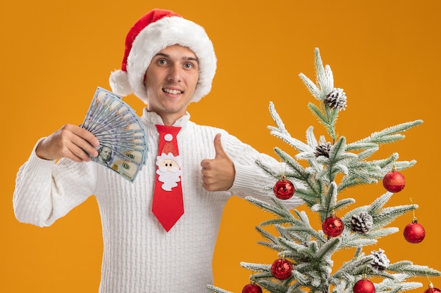 크리스마스 모자와 산타 클로스 넥타이를 입고 웃는 젊은 잘 생긴 남자가 돈을 들고 장식 된 크리스마스 트리 근처에 서있는 오렌지 벽에 고립 된 엄지 손가락을 보여주는