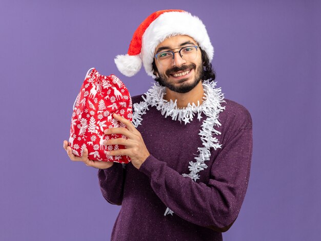 Smiling young handsome guy wearing christmas hat with garland on neck holding christmas bag isolated on blue background