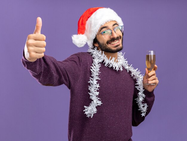 улыбающийся молодой красивый парень в новогодней шапке с гирляндой на шее, держащий бокал шампанского, показывая большой палец вверх изолированно на синей стене