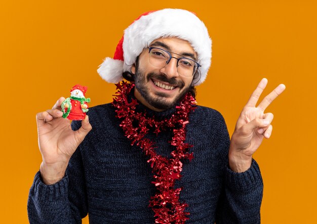 オレンジ色の壁に分離された平和のジェスチャーを示すおもちゃを持って首に花輪とクリスマスの帽子をかぶった笑顔の若いハンサムな男