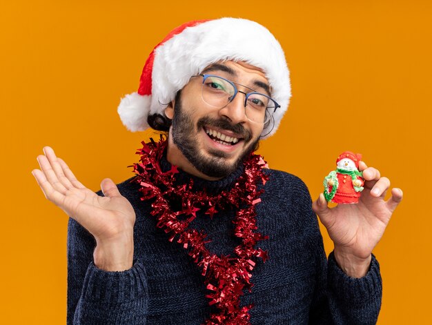 Улыбающийся молодой красивый парень в новогодней шапке с гирляндой на шее, держащий игрушку за руку, изолированную на оранжевом фоне