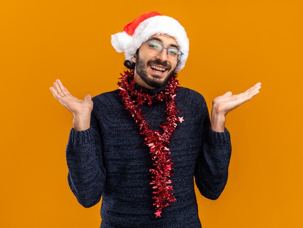 Smiling young handsome guy wearing christmas hat with garland on neck spreading hands isolated on orange background