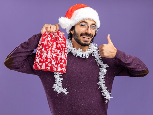 파란색 벽에 고립 엄지 손가락을 보여주는 크리스마스 가방을 들고 목에 갈 랜드와 함께 크리스마스 모자를 쓰고 웃는 젊은 잘 생긴 남자