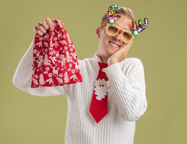 smiling young handsome guy wearing christmas novelty glasses and santa claus tie holding christmas sack looking at camera keeping hand on face isolated on olive green background