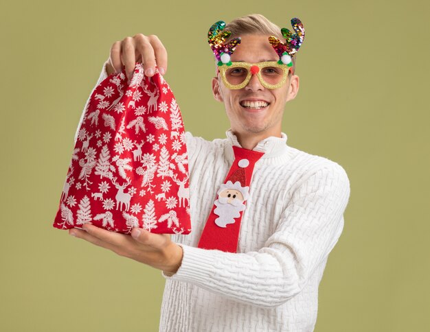 クリスマスノベルティメガネとサンタクロースのネクタイを身に着けている若いハンサムな男の笑顔は、オリーブグリーンの背景で隔離のカメラを見てカメラに向かってクリスマス袋を伸ばしています。