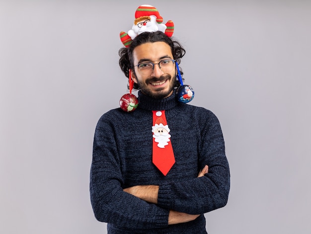 Free photo smiling young handsome guy wearing christmas tie with hair hoop hung christmas ball on ears crossing hands isolated on white wall