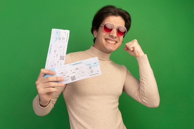 Free photo smiling young handsome guy wearing glasses holding tickets showing yes gesture isolated on green wall