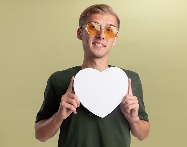 Sorridente giovane bel ragazzo che indossa la camicia verde con gli occhiali che tengono la scatola a forma di cuore isolato sulla parete verde oliva