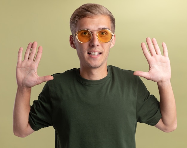 Smiling young handsome guy wearing green shirt with glasses raising hands isolated on olive green wall