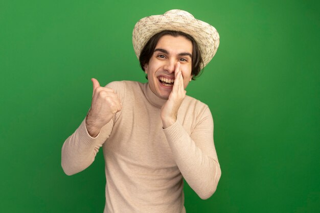Улыбающийся молодой красивый парень в шляпе указывает сбоку, положив руку вокруг рта, изолированного на зеленой стене с копией пространства