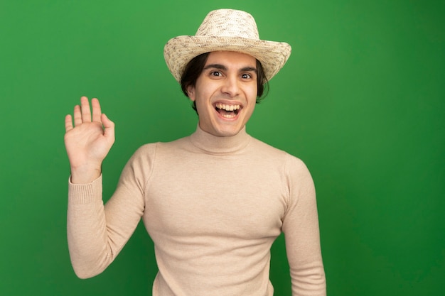 Улыбающийся молодой красивый парень в шляпе показывает приветственный жест изолирован на зеленой стене