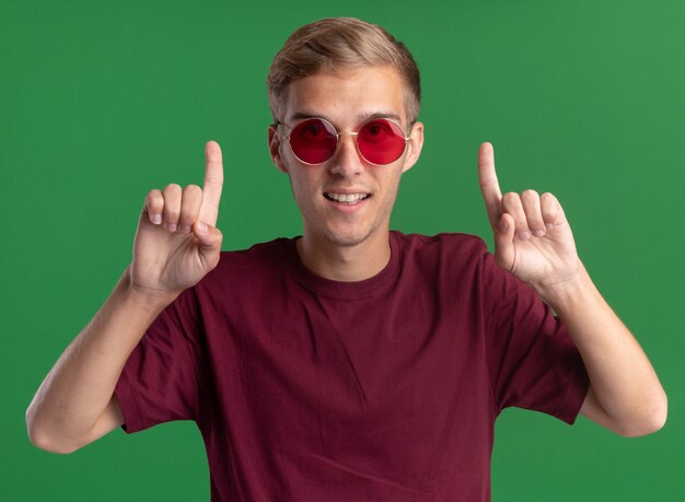 Smiling young handsome guy wearing red shirt and glasses points at up isolated on green wall