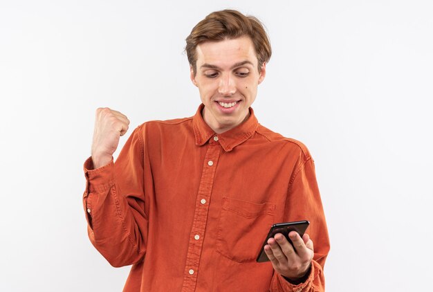 Smiling young handsome guy wearing red shirt holding and looking at phone showing yes gesture isolated on white wall