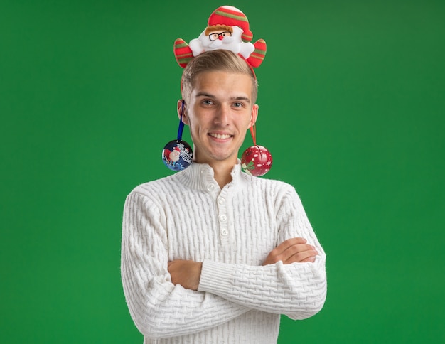 Free photo smiling young handsome guy wearing santa claus headband standing with closed posture looking at camera with christmas baubles hanging from ears isolated on green background with copy space