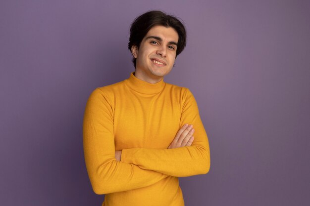 Улыбающийся молодой красивый парень в желтом свитере с высоким воротом, скрестив руки на фиолетовой стене с копией пространства