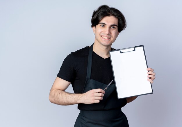 Smiling young handsome male barber in uniform holding clipboard with scissors isolated on white
