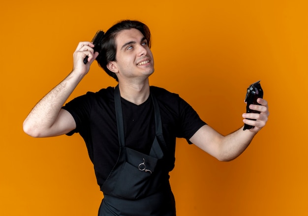 Smiling young handsome male barber in uniform holding hair clippers and combing hair isolated on orange