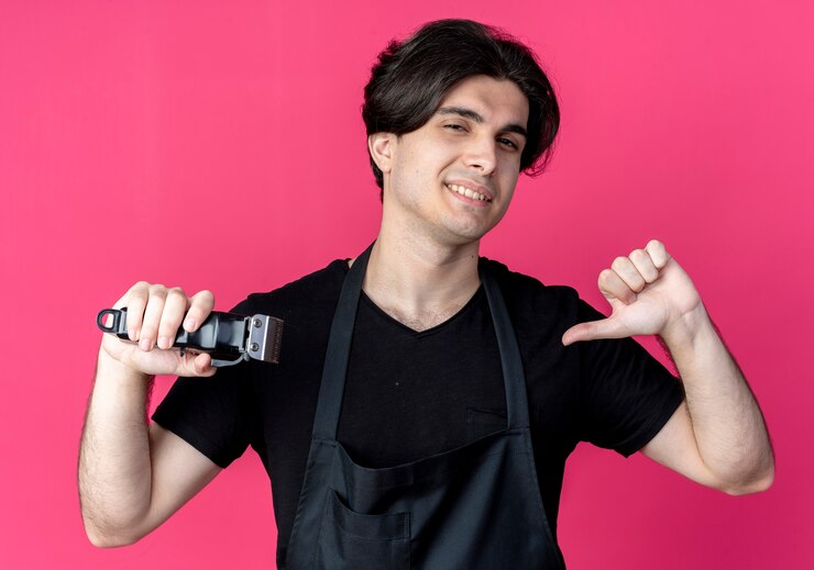मुफ़्त फोटो smiling young handsome male barber in uniform holding hair clippers and points himself isolated on pink
