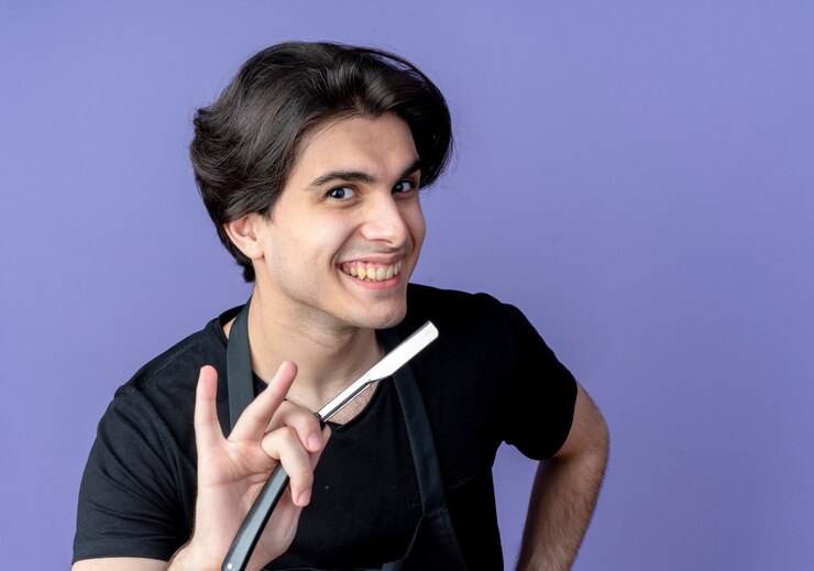 मुफ़्त फोटो smiling young handsome male barber in uniform holding out straight razor at camera isolated on blue