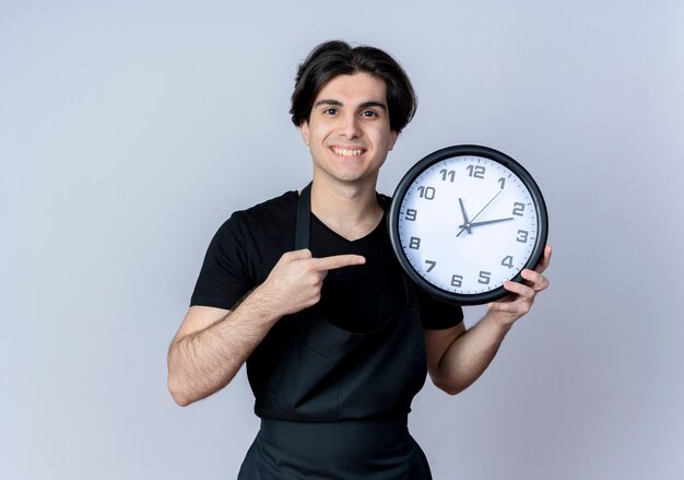 Smiling young handsome male barber in uniform holding and points at wall clock isolated on white