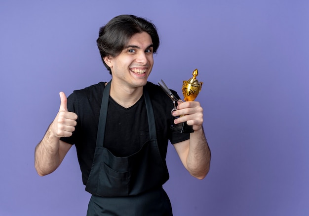 Free photo smiling young handsome male barber in uniform holding winner cup with scissors and his thumb up isolated on blue