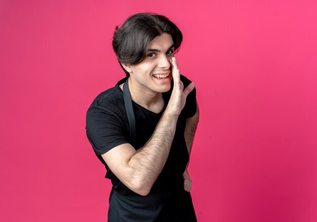 Smiling young handsome male barber in uniform whispers isolated on pink