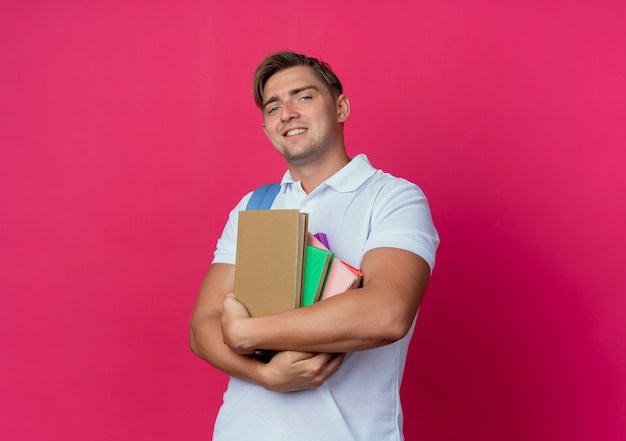 Smiling young handsome male student holding books isolated on pink