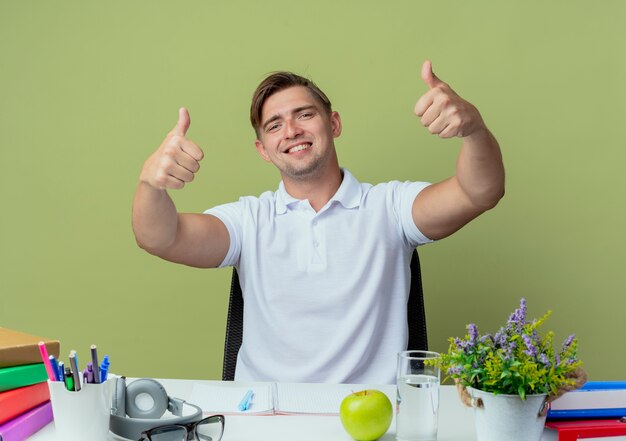 Smiling young handsome male student sitting at desk with school tools his thumbs up isolated on olive green