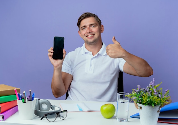 Smiling young handsome male student sitting at desk with school tools holding phone his thumb up isolated on blue