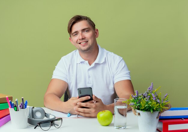 オリーブグリーンに電話を保持している学校のツールと机に座っている若いハンサムな男子生徒の笑顔