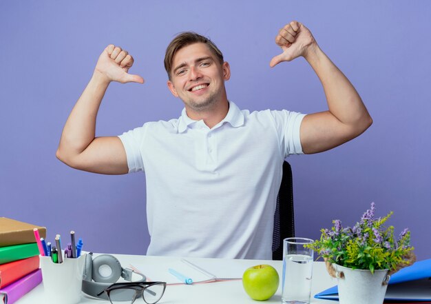 Smiling young handsome male student sitting at desk with school tools points himself isolated on blue