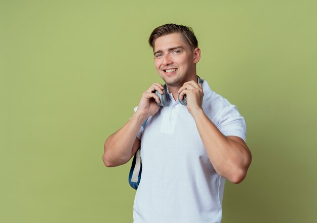 Smiling young handsome male student wearing back bag and headphones isolated on olive green