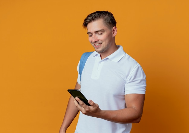 Smiling young handsome male student wearing back bag holding and looking at phone isolated on orange