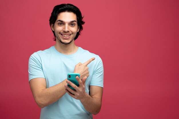 コピースペースと赤い背景で隔離の側を指しているカメラを見て携帯電話を保持している笑顔の若いハンサムな男