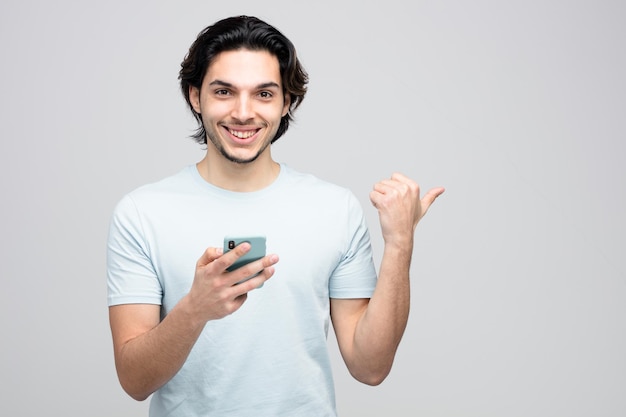 улыбающийся молодой красивый мужчина с мобильным телефоном, смотрящий в камеру, указывающую на сторону, изолированную на белом фоне с копировальным пространством