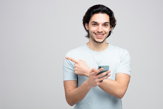 smiling young handsome man holding mobile phone looking at camera pointing to side isolated on white background with copy space