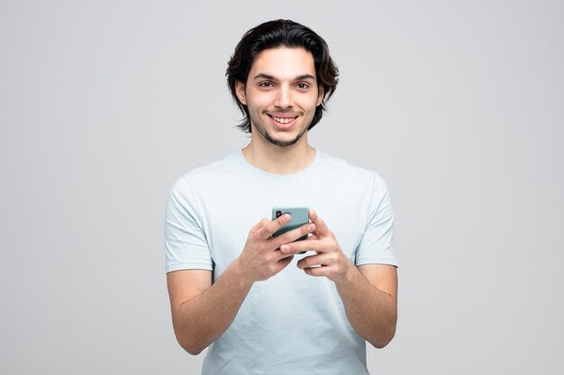 Улыбающийся молодой красивый мужчина держит мобильный телефон обеими руками, глядя на камеру на белом фоне