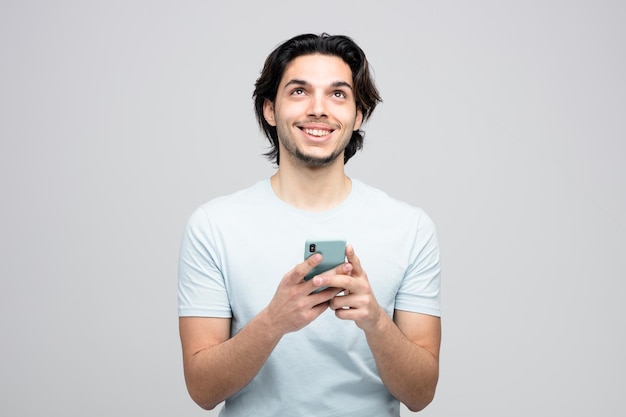 白い背景で隔離の見上げる両手で携帯電話を保持している若いハンサムな男の笑顔