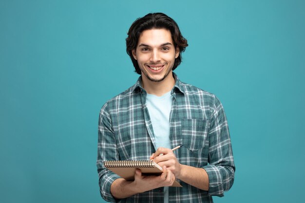 smiling young handsome man holding note pad and pencil looking at camera isolated on blue background