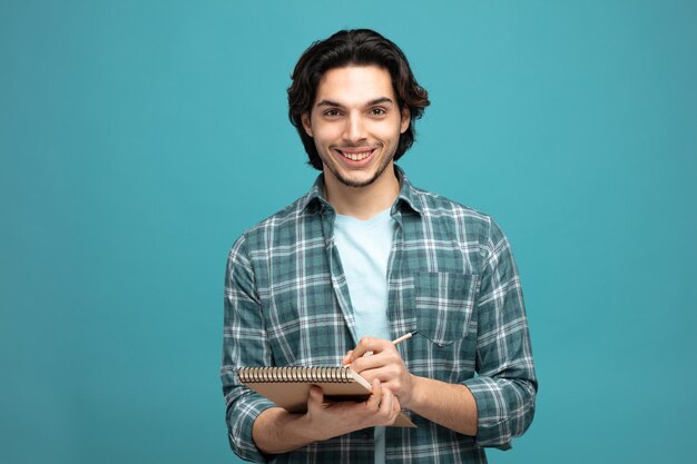 青い背景で隔離のカメラを見てメモ帳と鉛筆を保持している若いハンサムな男の笑顔