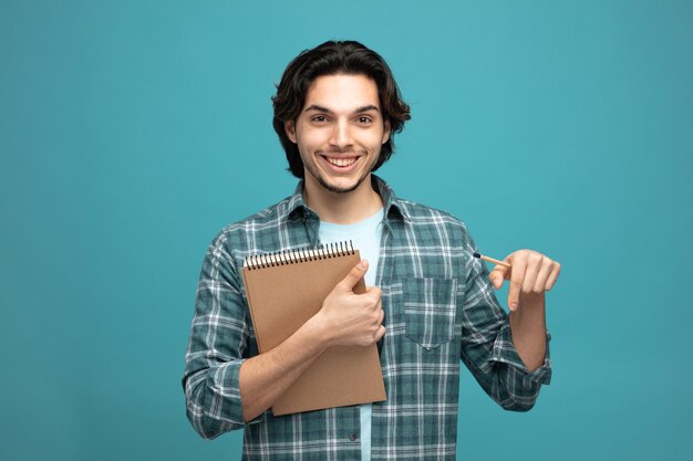 smiling young handsome man holding note pad and pencil looking at camera pointing down isolated on blue background