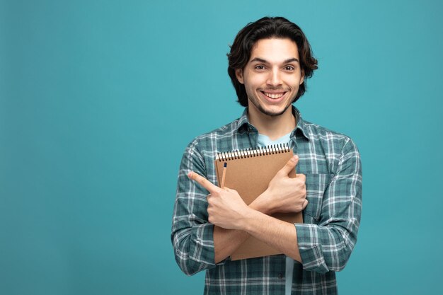 smiling young handsome man holding note pad and pencil looking at camera pointing to side isolated on blue background with copy space