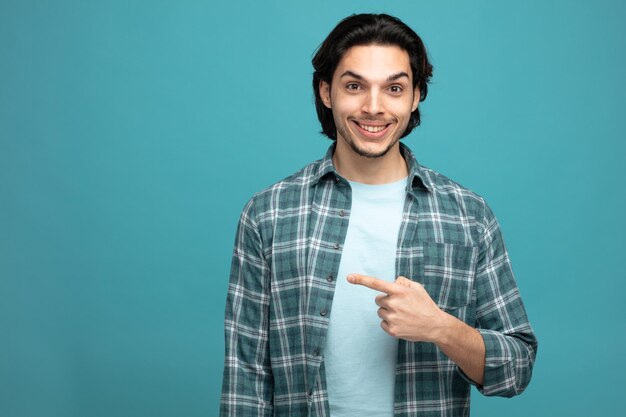 smiling young handsome man looking at camera pointing to side isolated on blue background with copy space