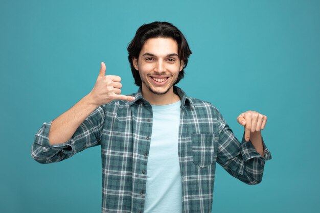 smiling young handsome man looking at camera showing call gesture pointing down isolated on blue background