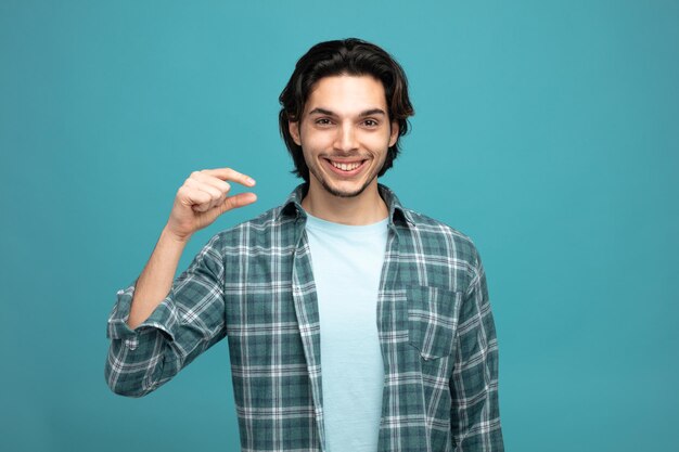 smiling young handsome man looking at camera showing small amount sign isolated on blue background