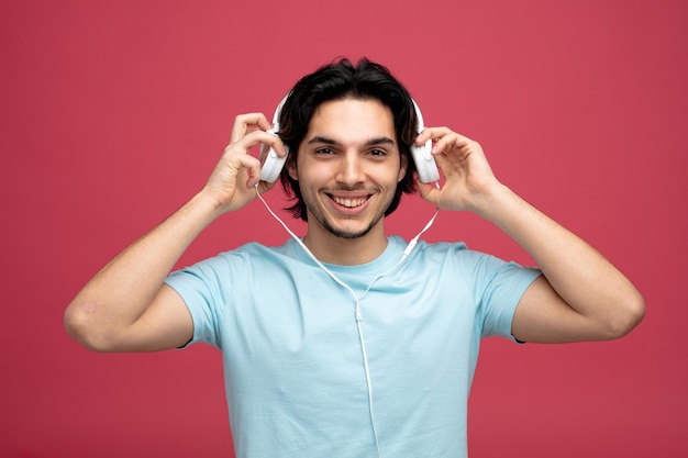 smiling young handsome man looking at camera taking headphones off isolated on red background