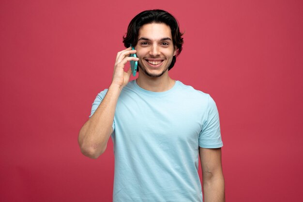 赤い背景で隔離の電話で話しているカメラを見て笑顔の若いハンサムな男